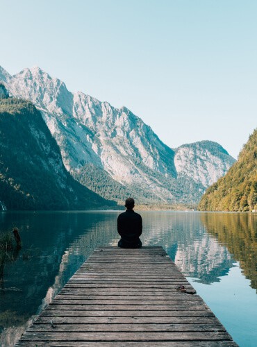 A person sits at the end of a wooden dock, facing a calm lake surrounded by majestic mountains under a clear blue sky. Like the tranquility found in motorcycle maintenance, the scene conveys peace and contemplation.