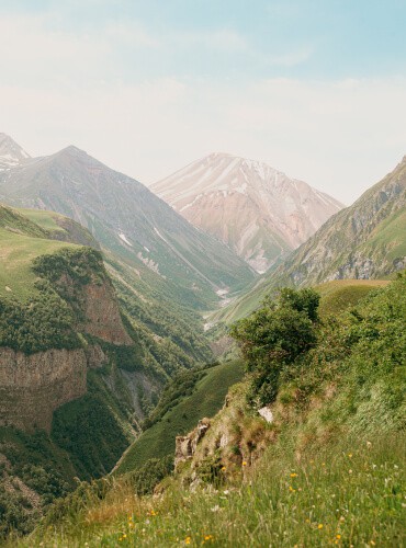 A scenic mountain landscape with lush green valleys and steep cliffs. In the background, tall peaks dusted with snow rise against a bright blue sky, evoking the adventurous spirit of motorcycle transport services. Small wildflowers are scattered across the foreground.