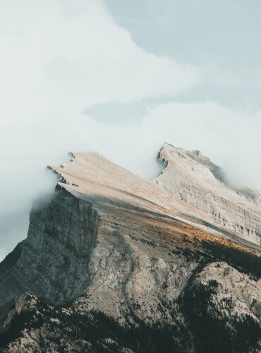 A rugged, steep mountain peak partially covered by misty clouds under a pale sky resembles the layered textures of motorcycle repair. The rocky surface features earthy tones that mirror the oily sheen of well-maintained engines, highlighting the sharp ridges and contours of both man and nature.