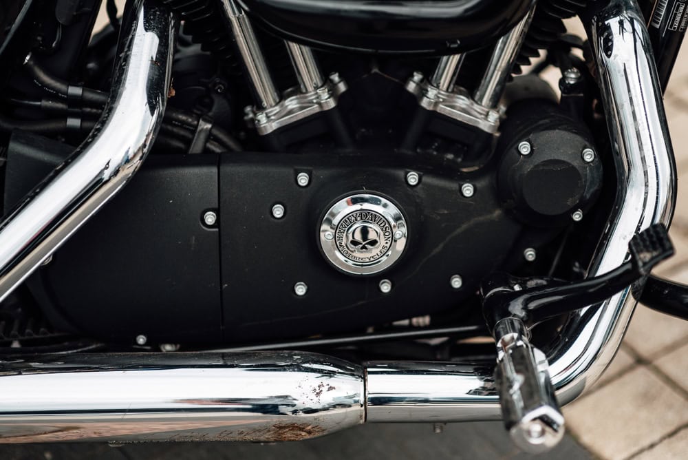 Close-up view of a meticulously detailed motorcycle engine with shiny chrome exhaust pipes. The engine features a round emblem in the center, surrounded by bolts, and is positioned on a sleek black casing. It's evident that expert motorcycle detailing enhances every gleaming inch.