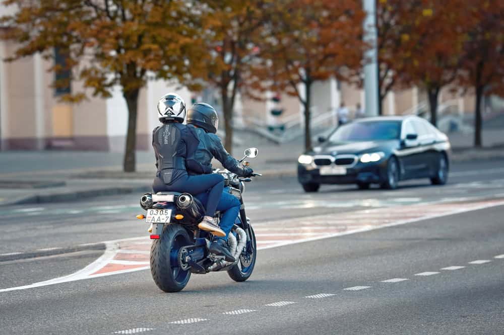 Two people wearing helmets ride a motorcycle on a city street, embodying the essence of meticulous motorcycle maintenance. In the background, a black car approaches. The street is lined with trees showcasing autumn foliage, and the buildings are slightly blurred.