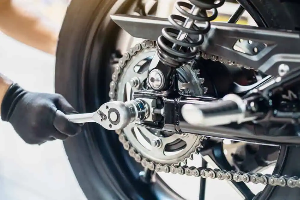 A person wearing black gloves uses a wrench to tighten a bolt on a motorcycle's rear wheel, showcasing the precision of motorcycle maintenance. The image focuses on the bike's suspension and chain mechanism.