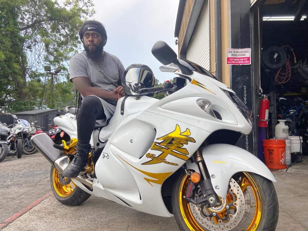 A man wearing a grey shirt and black pants sits on a white and gold motorcycle in a garage area with tires and tools around. The motorcycle has a sleek design with distinctive graphics. Other motorcycles are visible in the background.