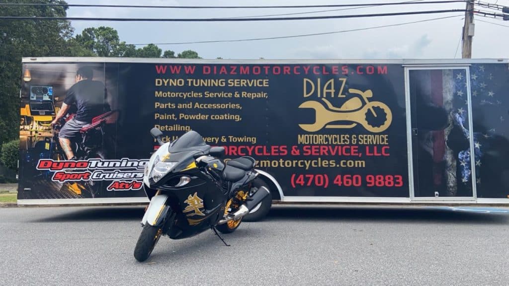 A sleek black motorcycle with gold accents is parked in front of a large trailer advertising "Diaz Motorcycles & Service." The trailer features graphics of motorcycles and services offered, such as dyno tuning, repairs, and detailing.