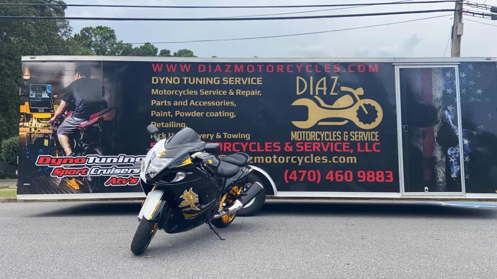 A sleek black motorcycle with gold accents is parked in front of a large trailer advertising "Diaz Motorcycles & Service." The trailer features graphics of motorcycles and services offered, such as dyno tuning, repairs, and detailing.