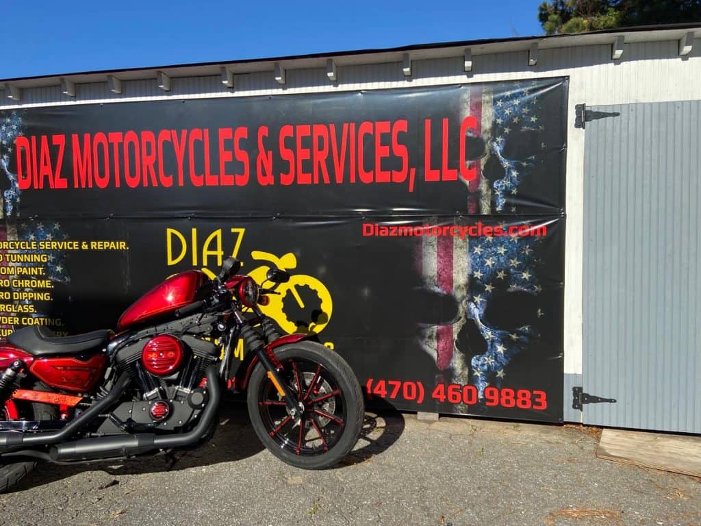 A red motorcycle is parked in front of a large sign for "Diaz Motorcycles & Services, LLC." The sign displays a logo with a skull and American flag design, along with contact information including a website and a phone number.