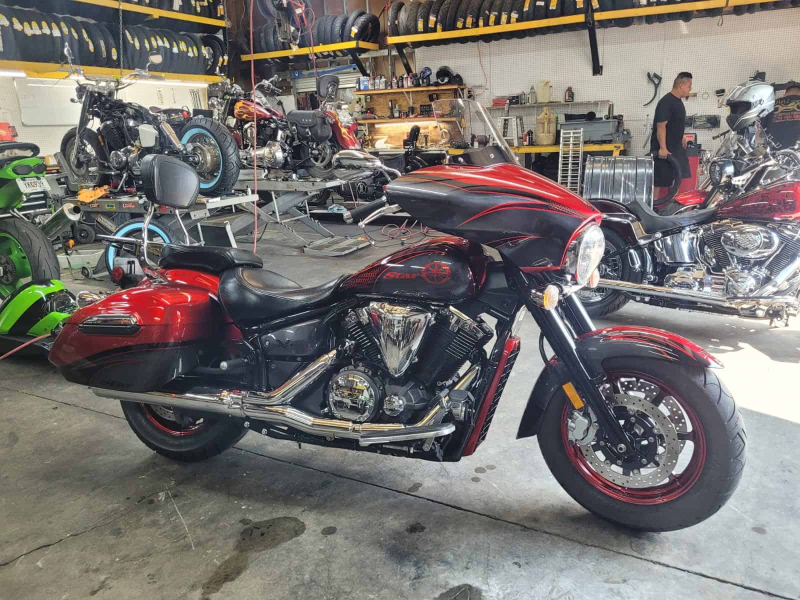 A red and black cruiser motorcycle in a workshop with various tools and equipment. Other motorcycles are visible in the background, and a person is standing near the back wall, possibly working on a bike. Rubber tires are stored on overhead shelves.