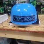 A blue helmet with "BX-FLYTY" printed on it, placed on a wooden shelf. In the background, there are boxes of air filters and various tools in a workshop setting.