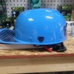 A shiny blue hard hat sitting on a wooden counter against a pegboard background, with black geometric logo on the side. Some tools and equipment are partially visible around it.