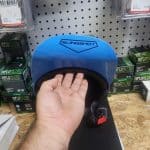 A person’s hand holding a blue helmet with "Slingshot" written on it, displayed against a pegboard wall. Various small box items are stacked around the helmet. The scene is indoors on a wooden counter.