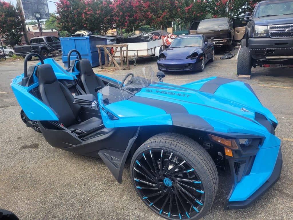 A bright blue Polaris Slingshot parked outdoors in a lot surrounded by other vehicles. The three-wheeled vehicle has unique, sporty design features with two seats and large, stylish wheels. In the background, various cars and a blue dumpster are visible.