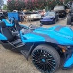 A bright blue Polaris Slingshot parked outdoors in a lot surrounded by other vehicles. The three-wheeled vehicle has unique, sporty design features with two seats and large, stylish wheels. In the background, various cars and a blue dumpster are visible.