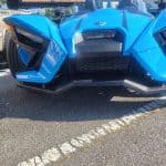 A bright blue, futuristic three-wheeled vehicle is parked in a lot. Its sleek design features sharp angles and an open cockpit. A sign advertising rooftop solar installation is visible in the background.
