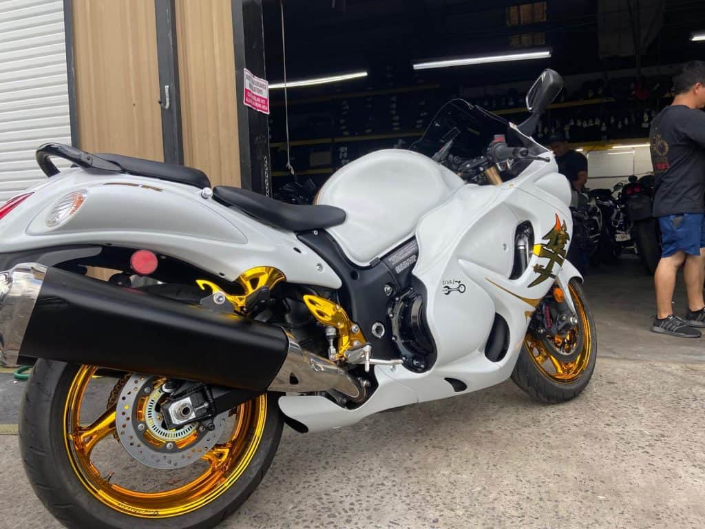 A white sport motorcycle with gold wheels and accents is parked outside a garage. A person is standing nearby in the background. The garage has a partially open door, revealing more motorcycles inside.