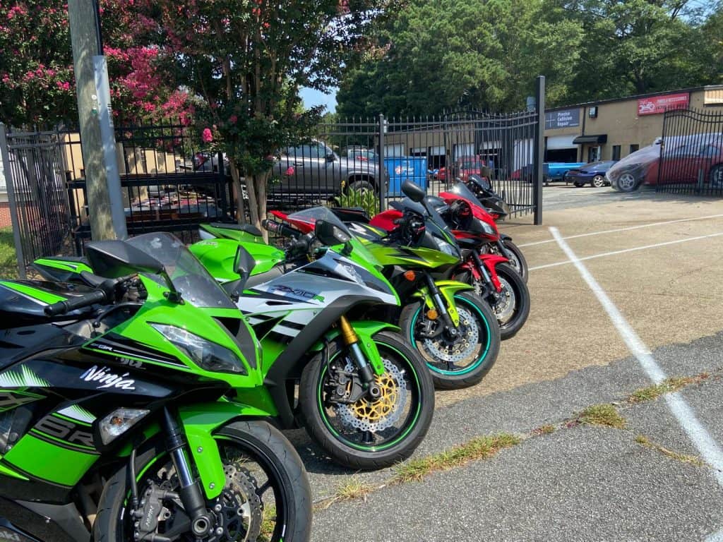 A row of brightly colored motorcycles, including green and red, parked in a lot. Trees and a fence are in the background, with a few cars and a building visible behind them. It's a sunny day.