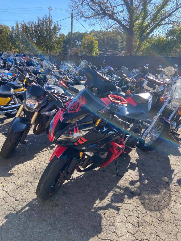 A parking lot filled with various motorcycles under the sunlight. Prominently in the foreground is a black and red motorcycle with a dragon design on its windshield. Trees and power lines are visible in the background.