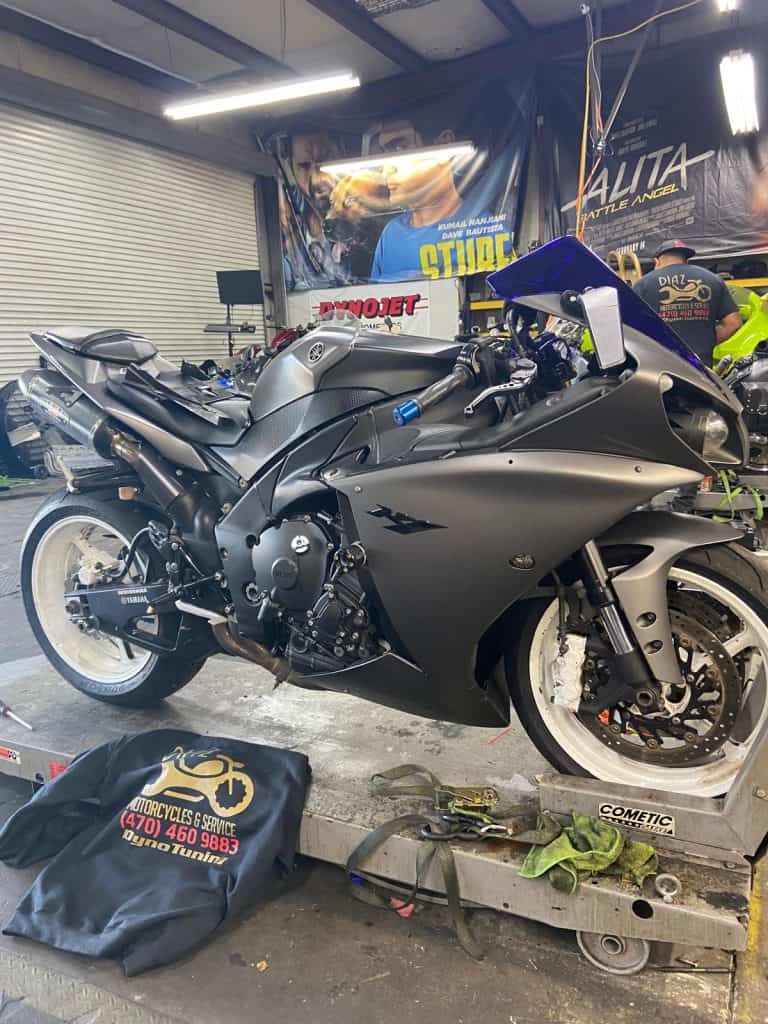 A sleek, matte black motorcycle is parked on a platform in a workshop. Surrounding the motorcycle are various tools and posters on the wall. A mat with a phone number is placed nearby. The space is well-lit with overhead lights.