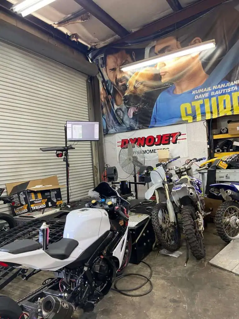 A workshop with various motorcycles, including a sport bike and dirt bike, on and near a Dynojet platform. A large poster with people and a dog hangs on the wall. There's equipment and boxes around the workspace.