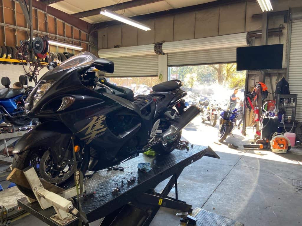 A black motorcycle is positioned on a lift inside a garage. Various tools and equipment are scattered around, and other motorcycles are visible in the background. Sunlight illuminates the open garage area.