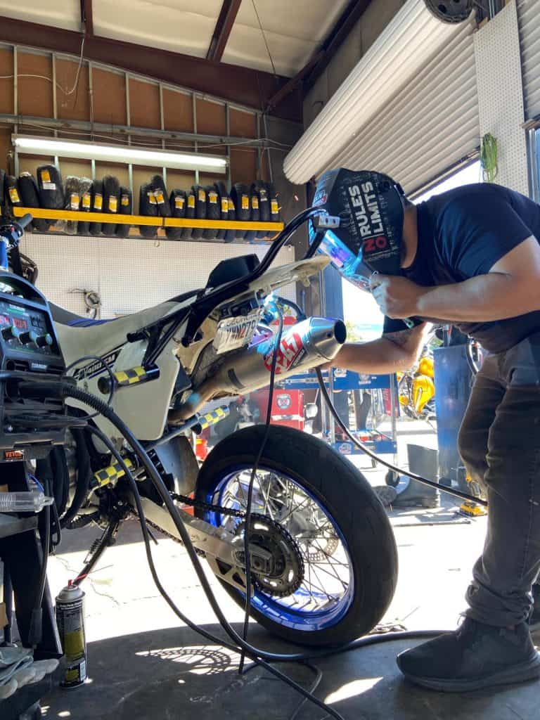 A person in a workshop is welding a motorcycle's exhaust. The workshop has tools on shelves, including oil containers. The welder wears a helmet that says "No Limits." Sunlight streams in through an open roll-up door.