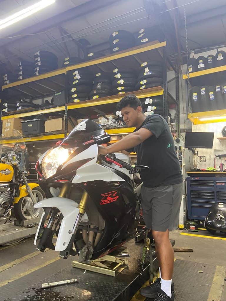 A person works on a motorcycle in a garage filled with various tools and tires. The motorcycle is elevated on a platform, and the person is focused on making adjustments. Shelves in the background hold motorcycle parts and equipment.