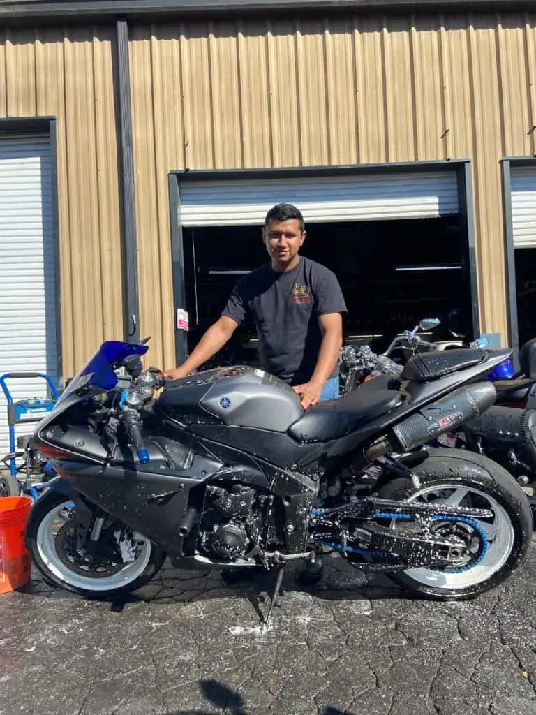 A person stands next to a gray motorcycle outside a building with partially open garage doors. The motorcycle is wet and soapy, indicating it's being washed. The ground is wet, and various equipment is visible in the background.