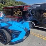 A blue three-wheeled vehicle is parked on the road next to a trailer featuring a motorcycle service advertisement. The trailer has graphics of a person tuning a motorcycle. The setting is outdoors on a sunny day.