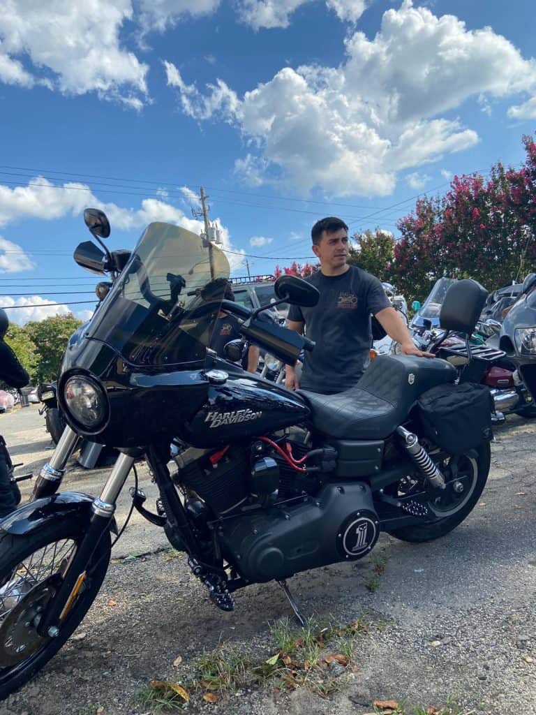 A man stands next to a parked black Harley-Davidson motorcycle on a sunny day. He's wearing a dark t-shirt, and there are more motorcycles in the background. The sky is partly cloudy, with trees lining the edge of the scene.