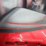 Close-up of a motorcycle tank with a sleek red and black paint job featuring a stylish red stripe design. The meticulous motorcycle detailing reveals its craftsmanship, while the background is blurred, capturing the essence of a bustling workshop setting.