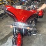 A custom red motorcycle with black decals is parked indoors, showcasing expert motorcycle detailing. The bike features a large front fairing, prominent headlights, and chrome details. The rider’s seat is designed for two, and a person's arm is visible in the background, interacting with the bike.