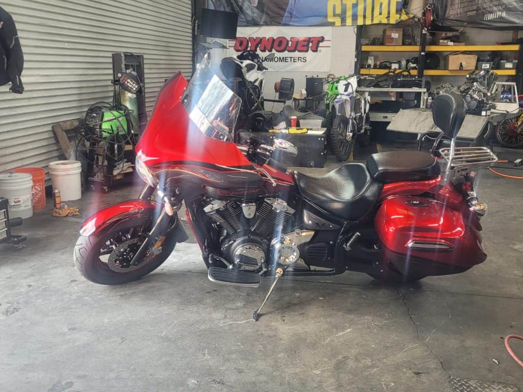 A red motorcycle with saddlebags, gleaming from recent detailing, is parked inside a garage. The bike, with a large front windscreen, stands on its kickstand amid various tools and equipment in the background.