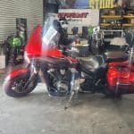 A red motorcycle with saddlebags, gleaming from recent detailing, is parked inside a garage. The bike, with a large front windscreen, stands on its kickstand amid various tools and equipment in the background.