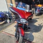 A bright red motorcycle, fresh from a meticulous detailing, is parked outside a garage with several other motorcycles. Two people stand nearby, one wearing a black shirt and the other in a blue shirt and cap, talking as the sun casts shadows on the concrete ground.