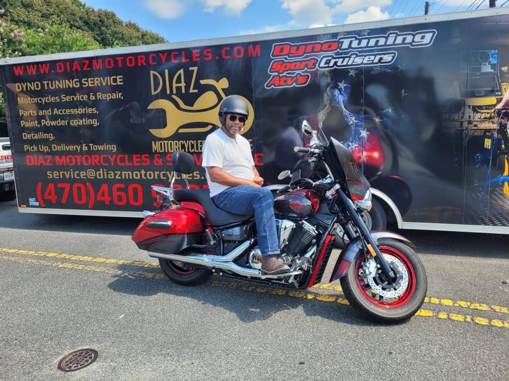 A person wearing a helmet sits on a red and black motorcycle, parked in front of a large motorcycle service trailer adorned with bold text and logos, ready for some expert motorcycle detailing on this sunny day.