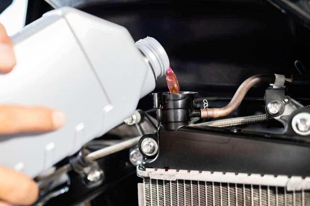 A person pours pink engine coolant from a plastic bottle into a car’s radiator under the hood. The radiator cap is open, and part of the radiator and engine components are visible.