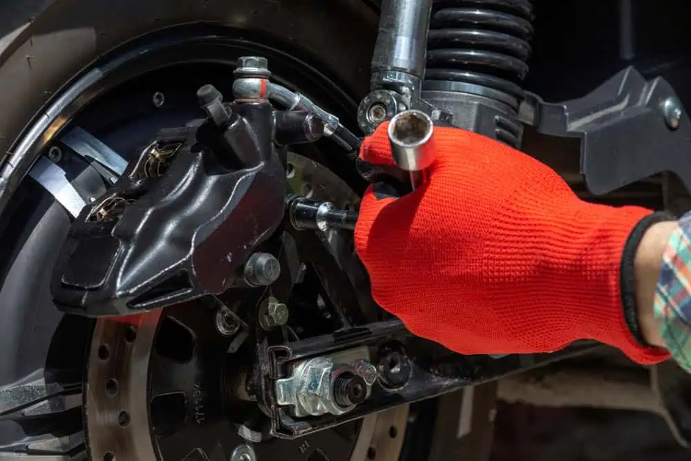 A person wearing a red glove uses a socket wrench to work on the rear disc brake of a motorcycle.