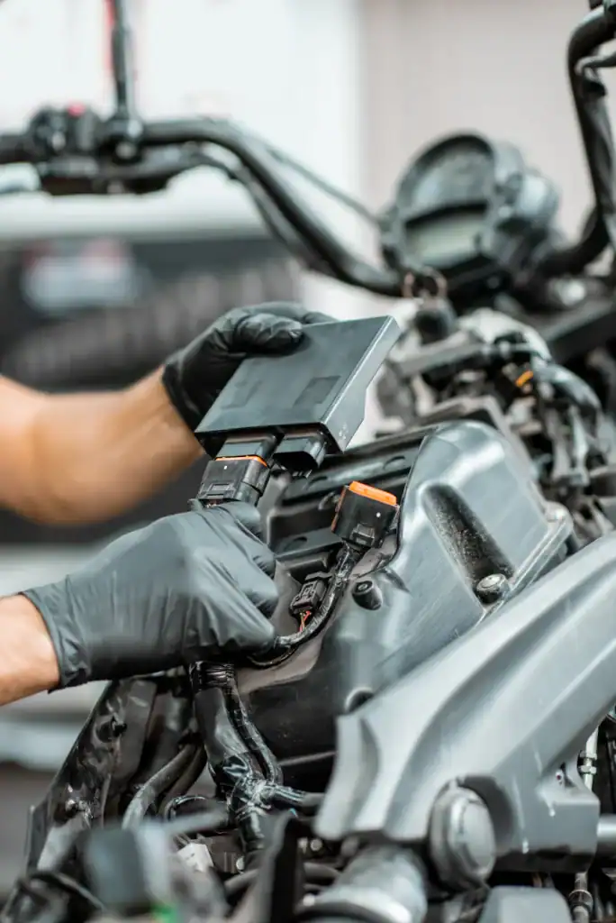 A person wearing black gloves uses diagnostic equipment to check the engine of a motorcycle, focusing on the exposed wiring and components near the handlebars.
