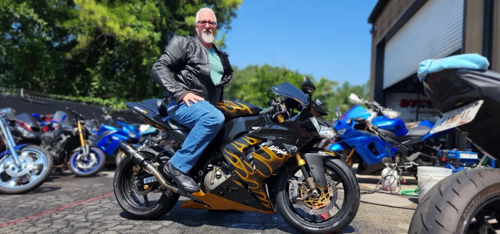 Bearded man in a leather jacket sitting on a black Kawasaki Ninja with bold gold flame paintwork outside the shop bays.