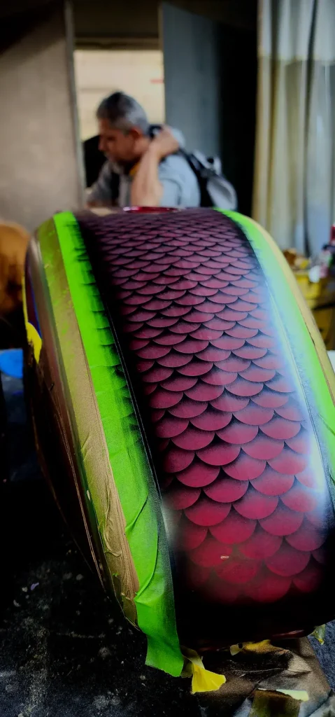 Close-up of a fuel tank freshly airbrushed with a deep red dragon-scale pattern, edges still wrapped in green masking tape, with a man photographing behind.