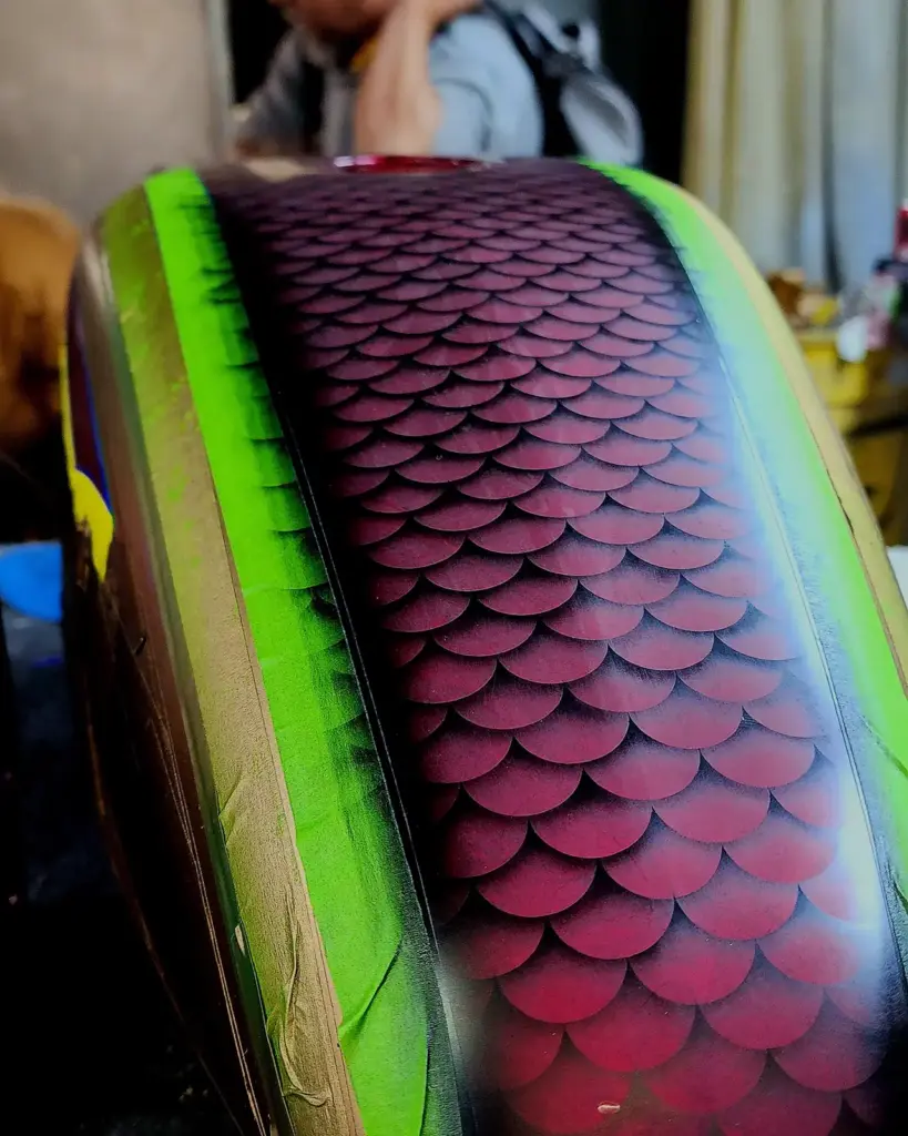 A close-up shows a painted object with red scales, green edges, and a blurred person in the background.