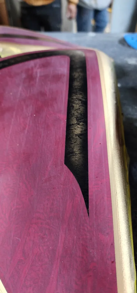 Extreme close-up of a magenta/candy purple painted panel with a black flame-like graphic and a gold pinstripe edge, showing swirl texture in the base coat.
