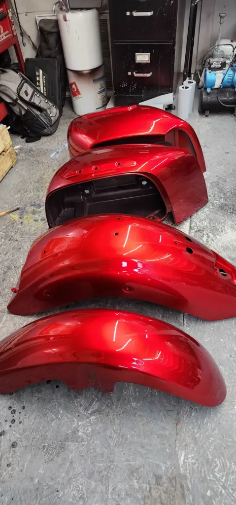 Four shiny red motorcycle fenders rest on a shop floor, surrounded by tools and workshop equipment.