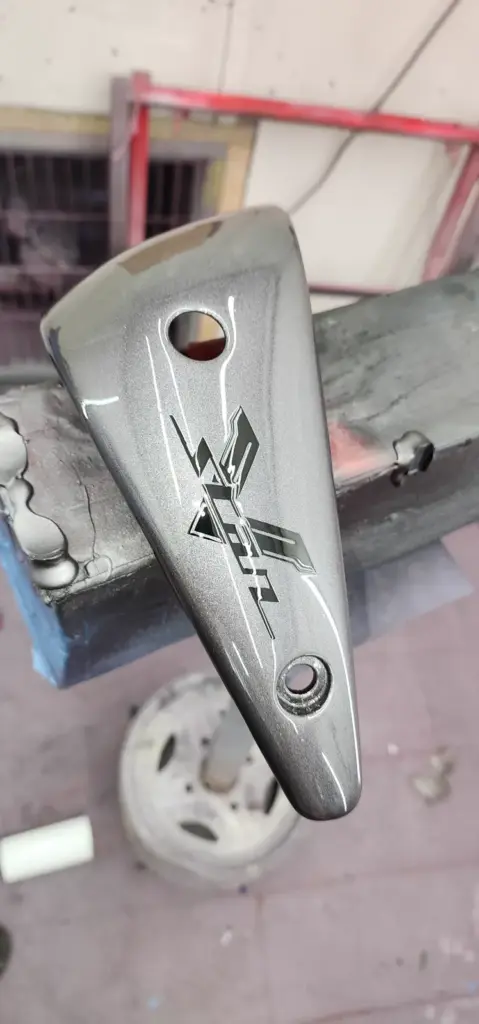 A metallic grey motorcycle part featuring a black “X” logo is displayed on a stand in a workshop.