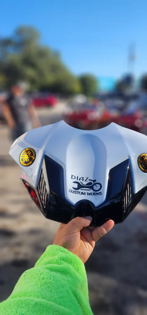 Hand holds up the finished white and black Yamaha tank cover with gold emblems and Diaz Custom Works logo against a blue sky outdoors.