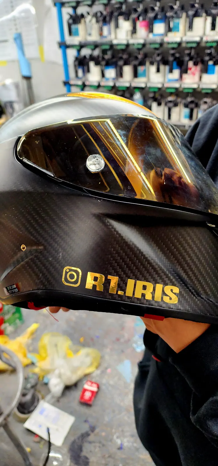 best custom paint Douglasville — hands holding a carbon-fiber helmet with a gold visor by the paint tint rack