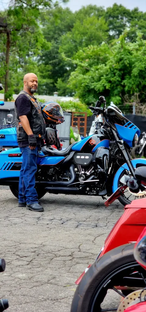 Side view of a custom blue and orange Harley bagger with matching blue trike behind it, owner in a leather vest standing alongside.