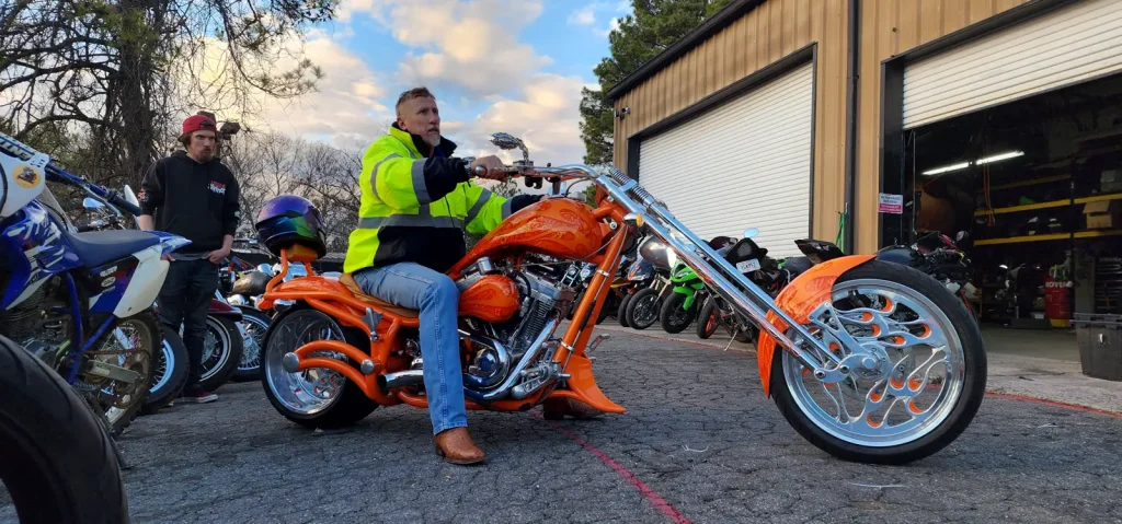 Wider angle of the same orange custom chopper with rider seated, dirt bike and shop bikes lined up alongside at dusk.