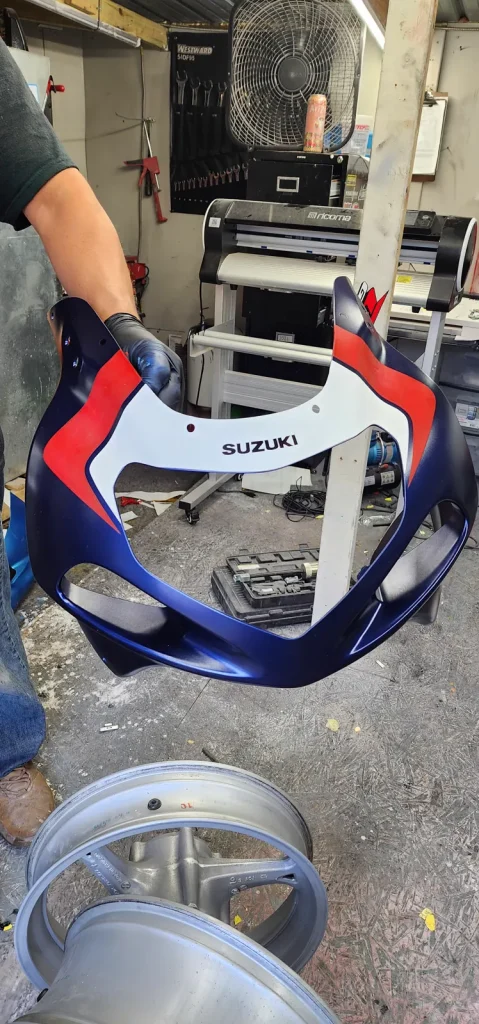 A worker in nitrile gloves holds up a freshly painted navy blue, red and white Suzuki upper fairing/nose cowl in the shop.