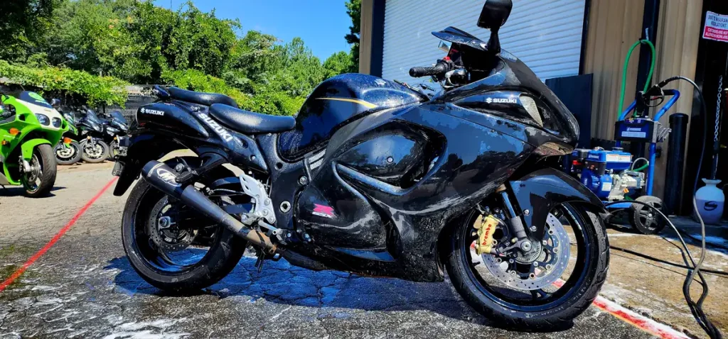 Black Suzuki Hayabusa standing on wet, soapy pavement mid-wash outside the shop bay, pressure washer and hose reel to the right.