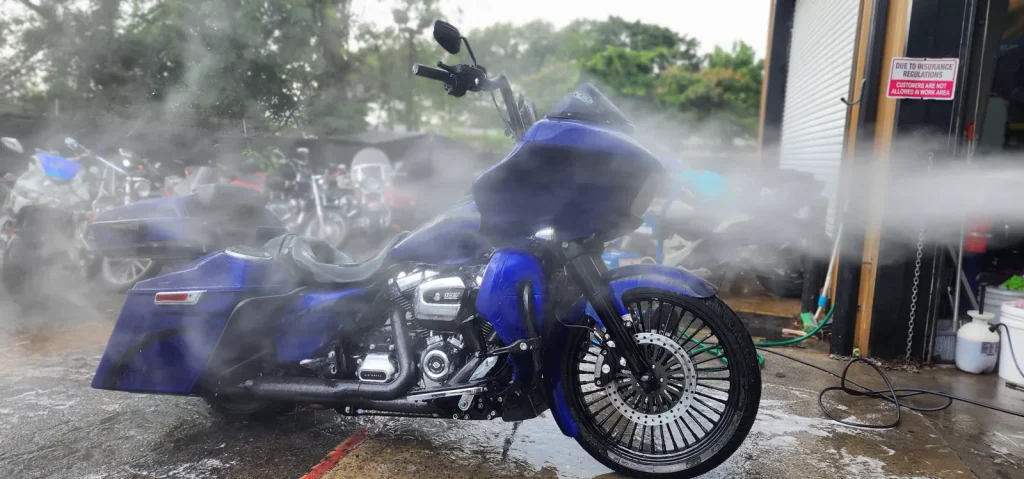 Blue Harley Road Glide being pressure-washed outside the shop, spray mist surrounding the bike and water running across the pavement.